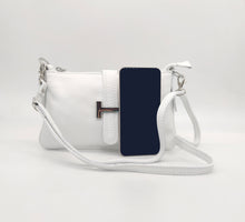 Cargar imagen en el visor de la galería, Bolso de piel blanco, con hebilla en plateado de adorno. Cierre con clip imantado en la parte trasera, y con cremallera. Lleva asa cortita para llevar al hombro, y asa larga austable para llevar colgado. Adornos en color plata. Interior forrado textil, con bolsillo plano con cremallera. LaNievina. Vista con móvil para referencia de tamaño