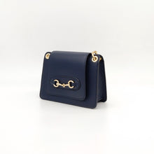Cargar imagen en el visor de la galería, Bolso de piel con cadena Géminis, de LaNievina, con adorno metálico en el frontal y asa de cadena con hombrera, vista frontal, color azul marino, cerrado, con adorno metálico dorado