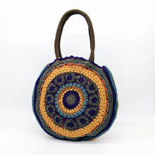Cargar imagen en el visor de la galería, Bolso redondo de colores de crochet en lana acrílica. Dos asas cortas y otra larga de lana trenzada, de LaNievina. Vista frontal modelo en azul y verde.